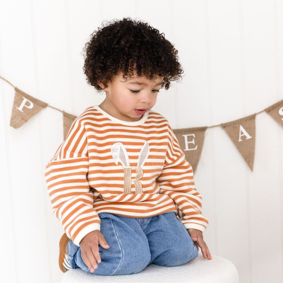 Child wearing a striped sweater with a bunny design, sitting on a white surface with a decorative banner in the background.