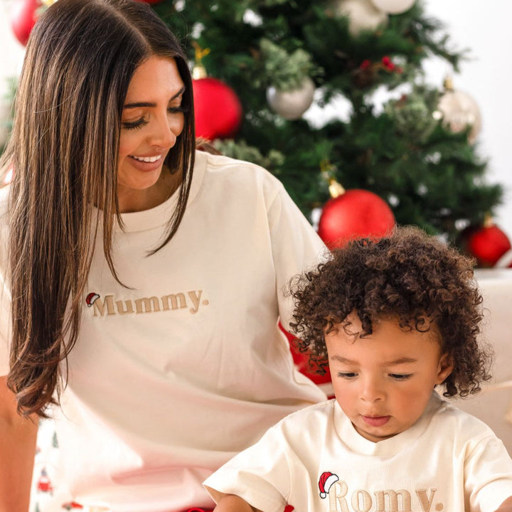 Woman and child sitting in front of a decorated Christmas tree wearing matching 'Mummy' shirts.