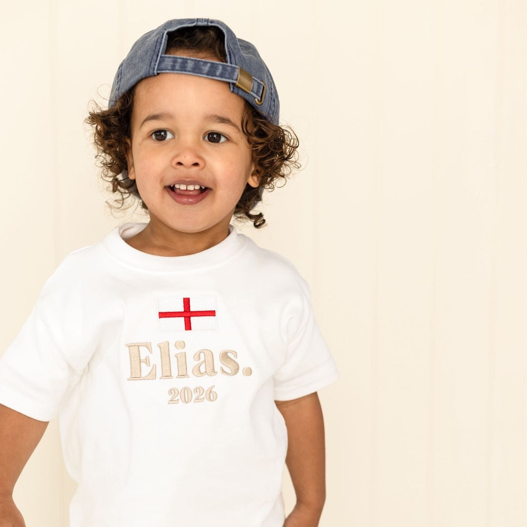 Child wearing a white t-shirt with 'Elias 2026' text and a blue cap on a beige background